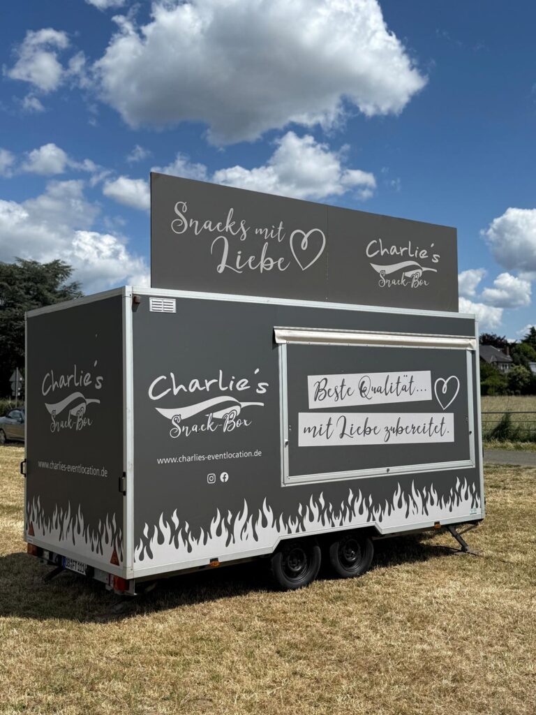 Grauer Imbisswagen mit weißer Schrift steht auf einer Wiese unter blauem Himmel