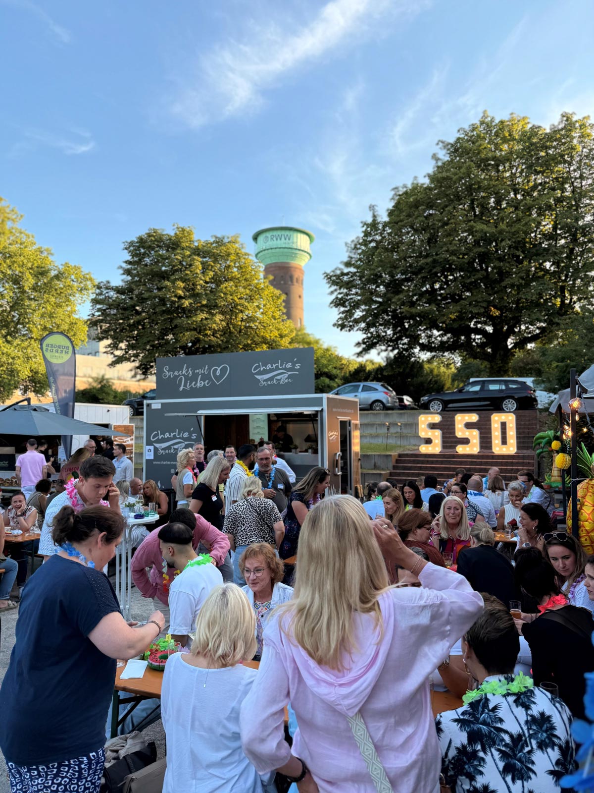Menschen genießen fröhlich ein Straßenfest mit Foodtrucks, Bäumen und Wasserturm im Hintergrund