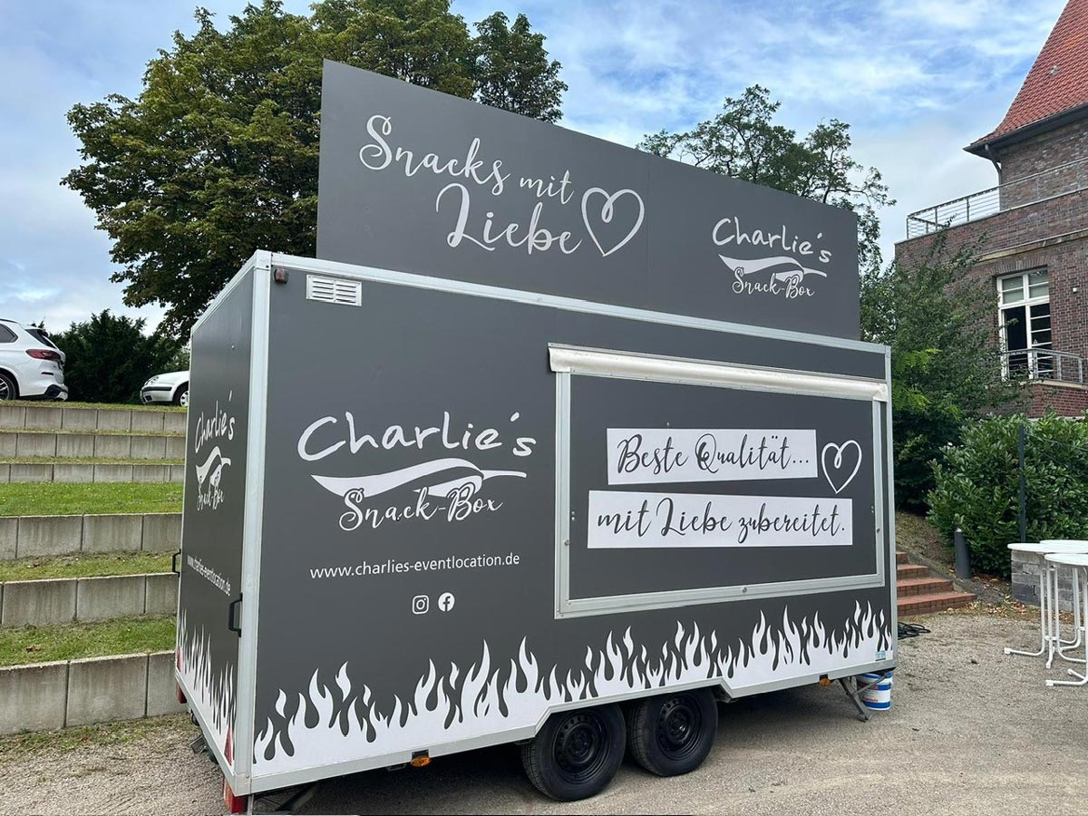 Ein grau-weißer Foodtruck mit Flammenmuster und großen Schildern steht draußen