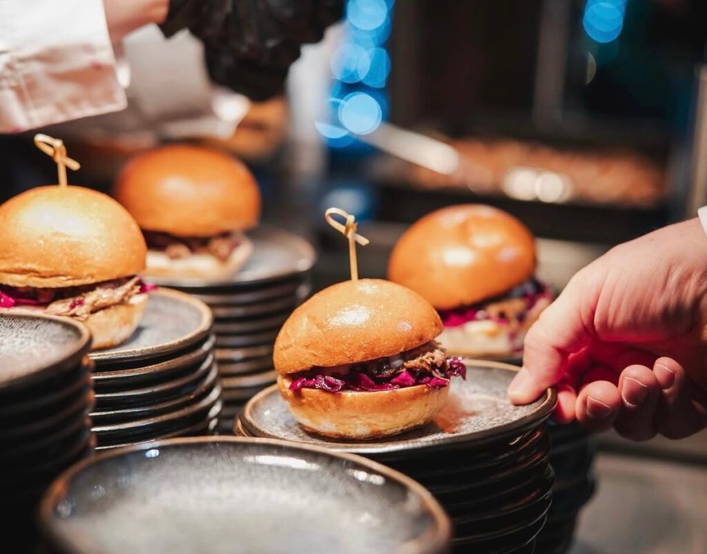 Eine Hand serviert Mini Burger mit Rotkohl auf gestapelten grauen Tellern